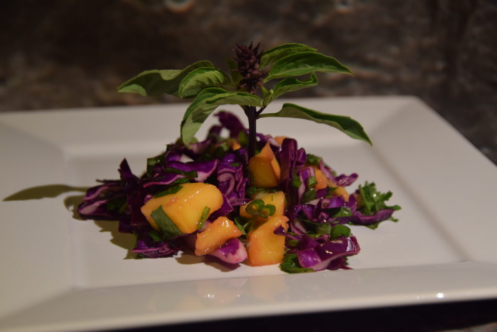 Orange mango, purple and green Thai basil, purple cabbage, and other green herbs in a slaw on a white plate, with a "tree" sprig of Thai basil in the center.