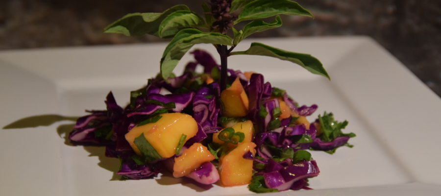 Orange mango, purple and green Thai basil, purple cabbage, and other green herbs in a slaw on a white plate, with a "tree" sprig of Thai basil in the center.