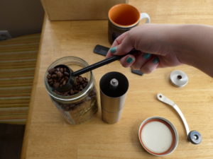 Scooping coffee beans into a JavaPresse coffee grinder, which is open, the crank on the table next to the mason jar lid, a mug in the background.