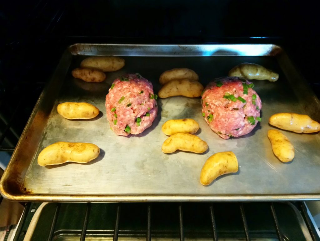 Image shows a silver sheet tray pan in the oven, with two uncooked meatloaves surrounded by yellow fingerling potatoes.