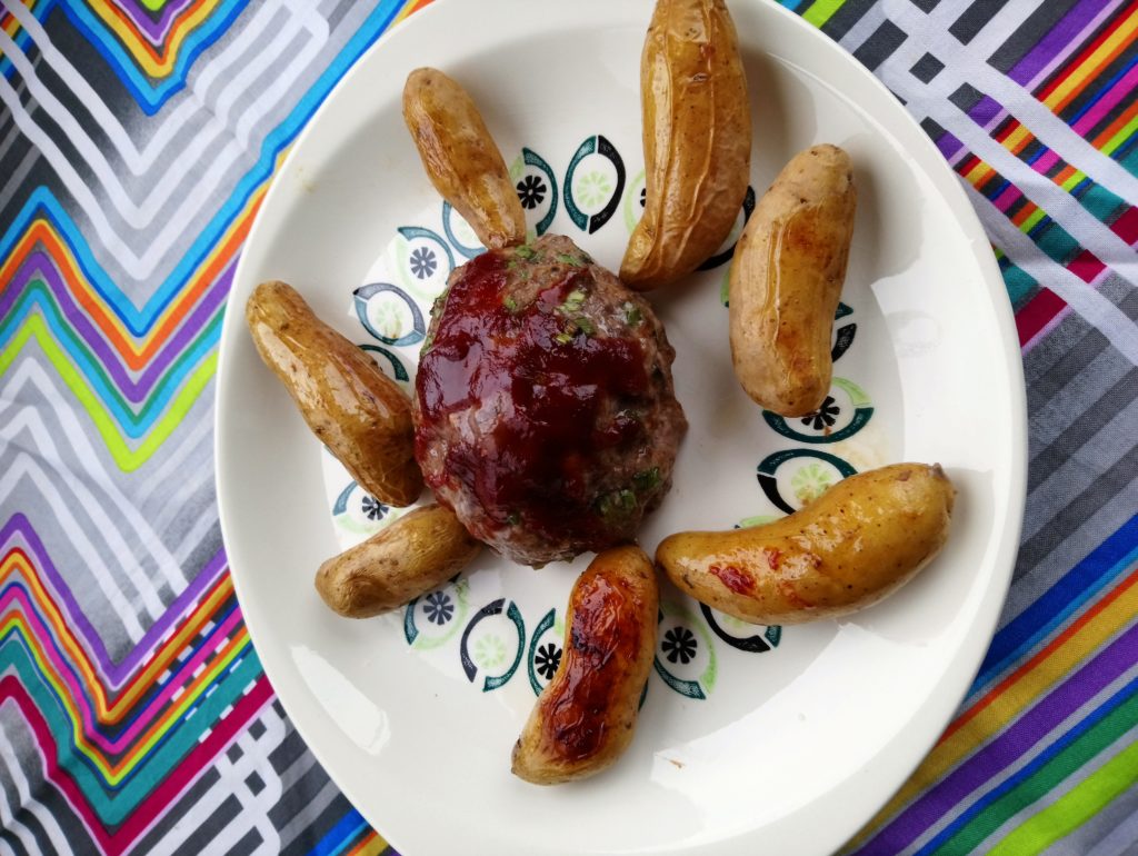 Image shows a platter with a green and black pattern, set on a multi-color zig-zag background. The platter holds a small meatloaf with a reddish sauce on top, surrounded by roasted fingerling potatoes.